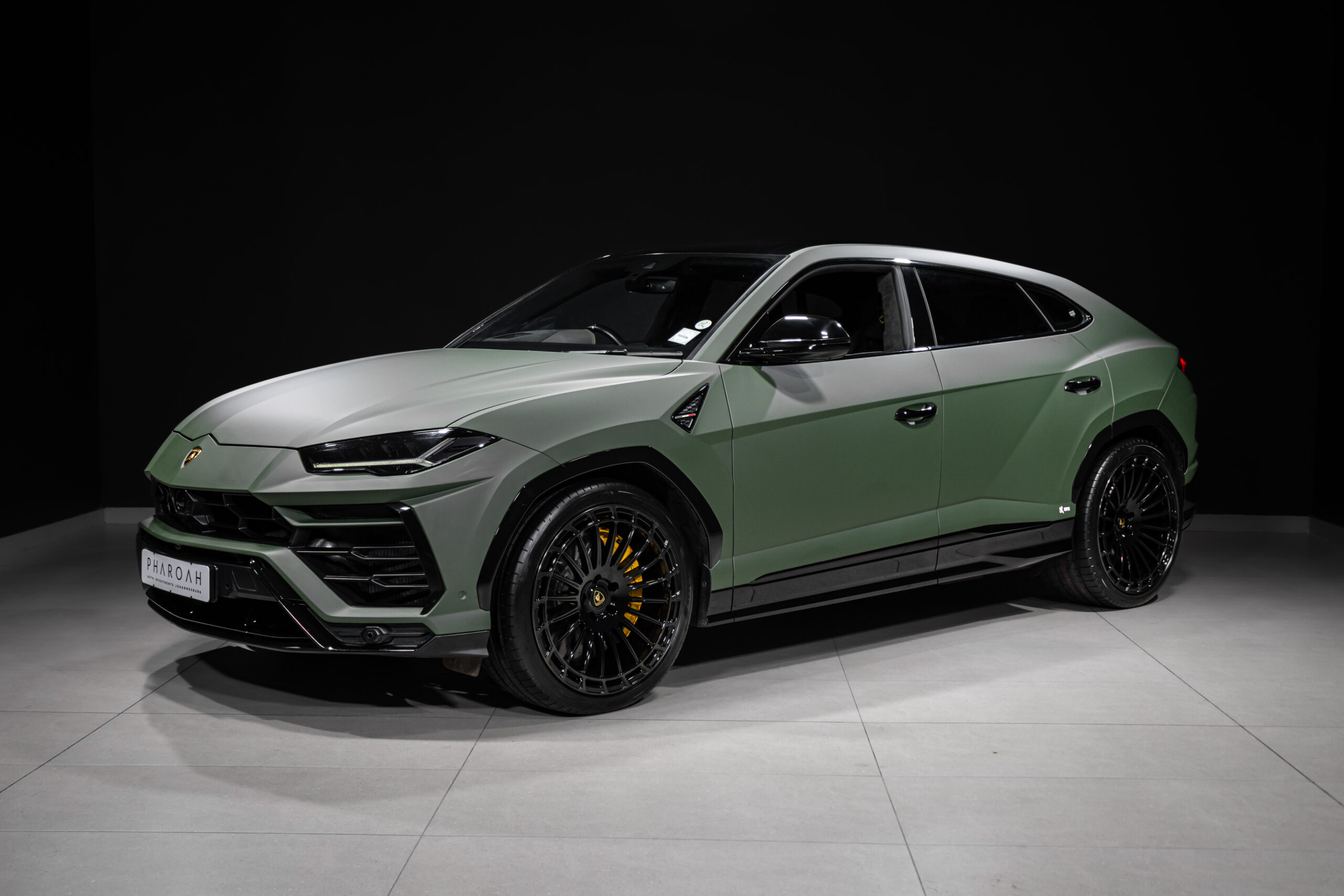 Three-quarter front-left view of a matte-green Lamborghini SUV on a showroom floor, with black wheels and dark background.