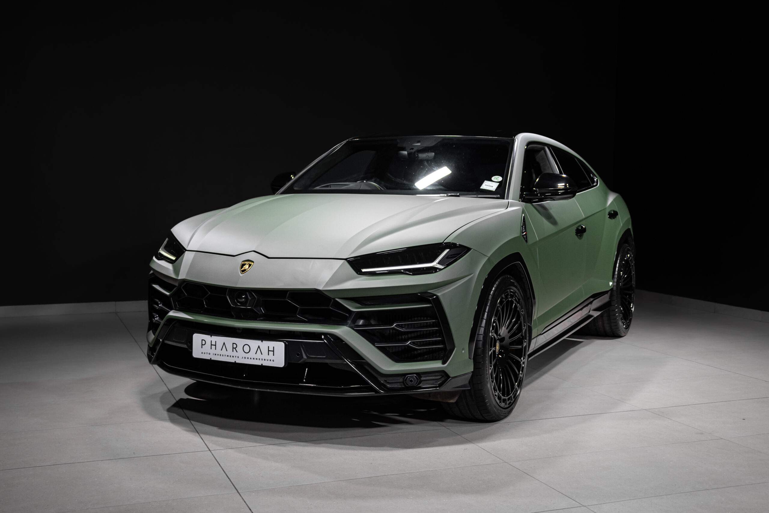 Front three-quarter view of a matte green Lamborghini Urus in a dark showroom with glossy black accents and large alloy wheel