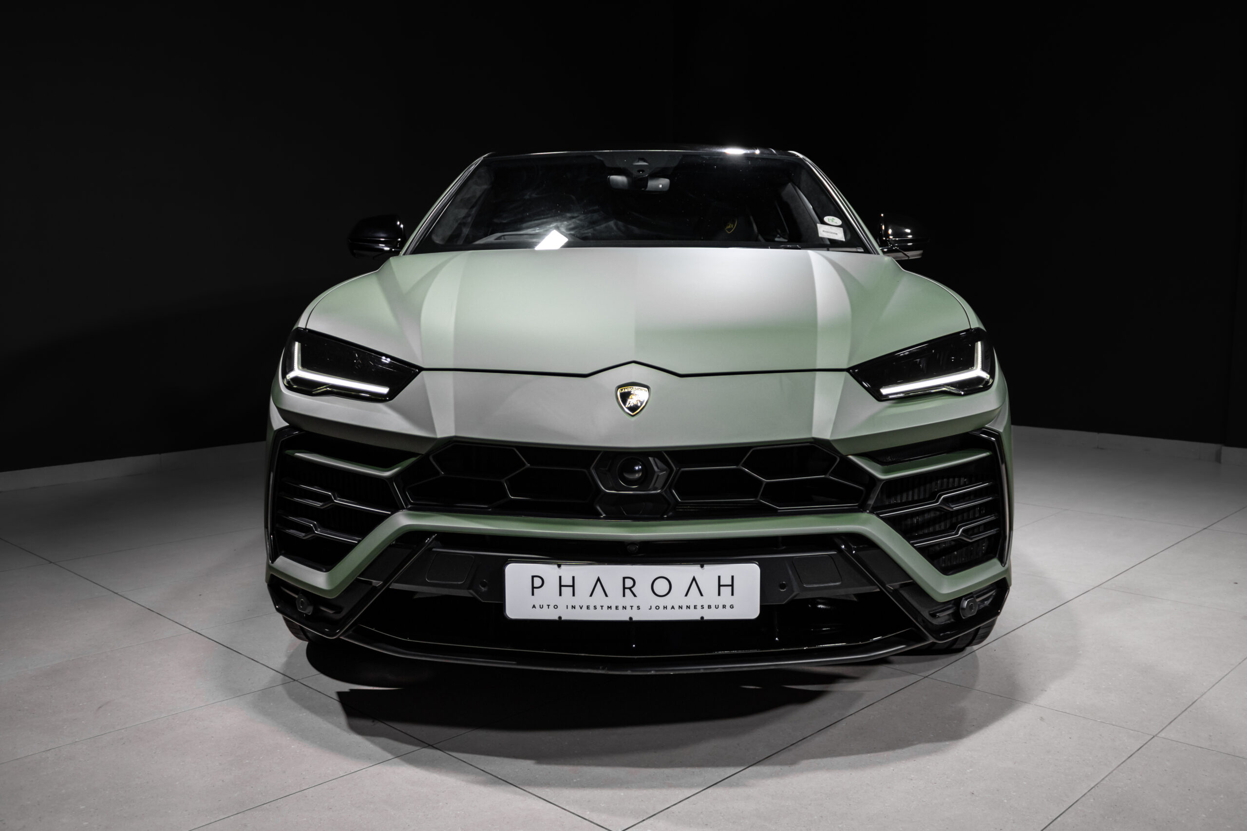 Front view of a pale green Lamborghini in a dark showroom, with distinctive angular headlights and a central badge above the