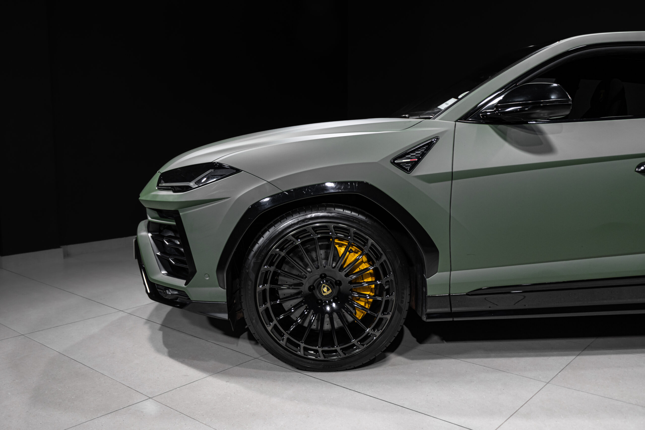 Close-up of the front left side of a matte-green luxury SUV, highlighting the large black multi-spoke wheel with yellow brake
