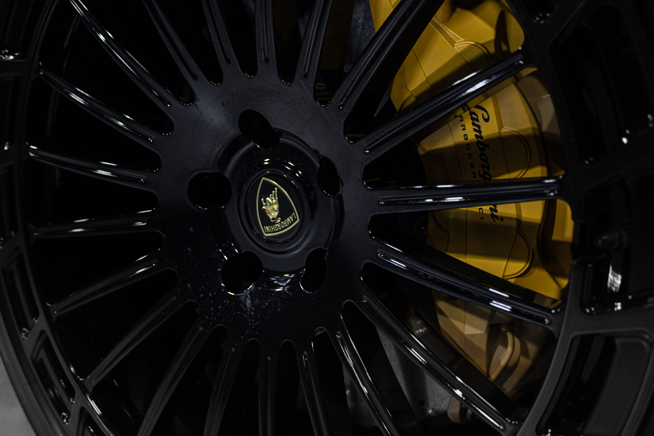 Close-up of a black gloss alloy wheel with a yellow brake caliper and the Lamborghini center cap logo visible.