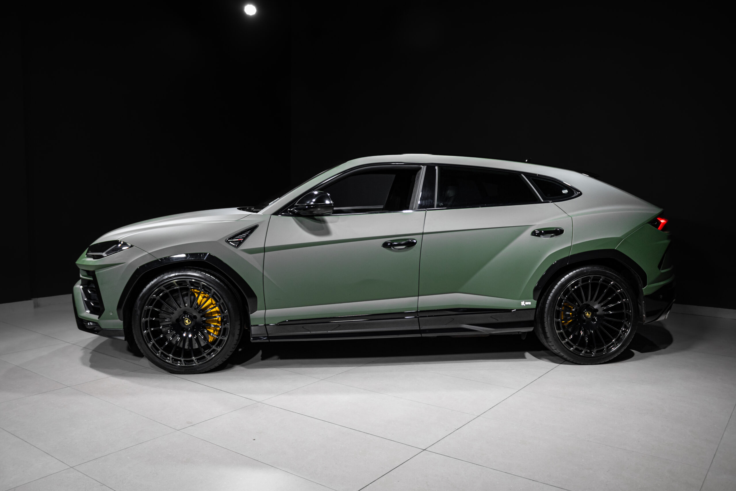Side profile of a green luxury SUV with large black alloy wheels and yellow brake calipers in a dark showroom backdrop