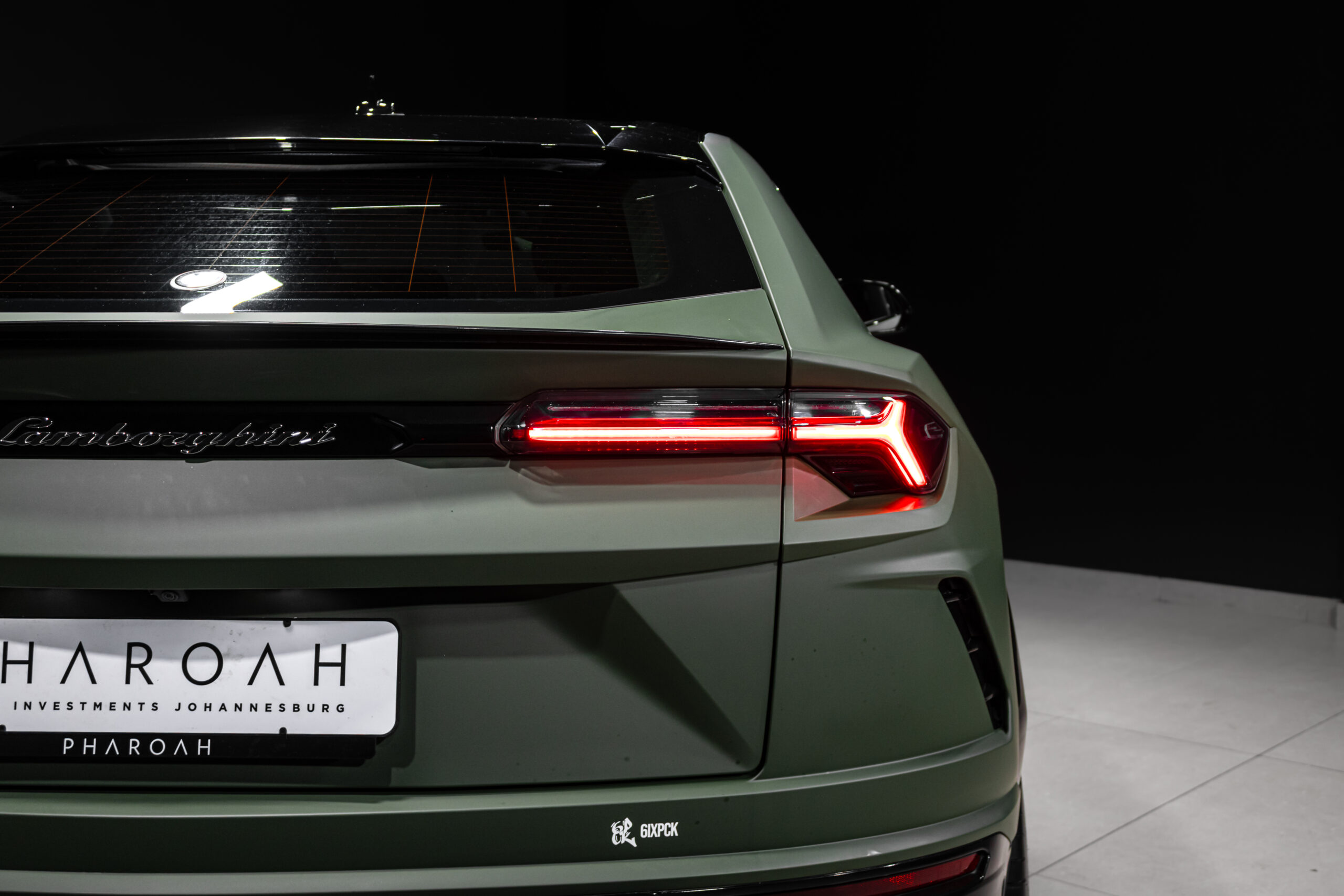 Rear view of a green Lamborghini in a dark showroom, with illuminated red taillights and the 'Lamborghini' script badge visib