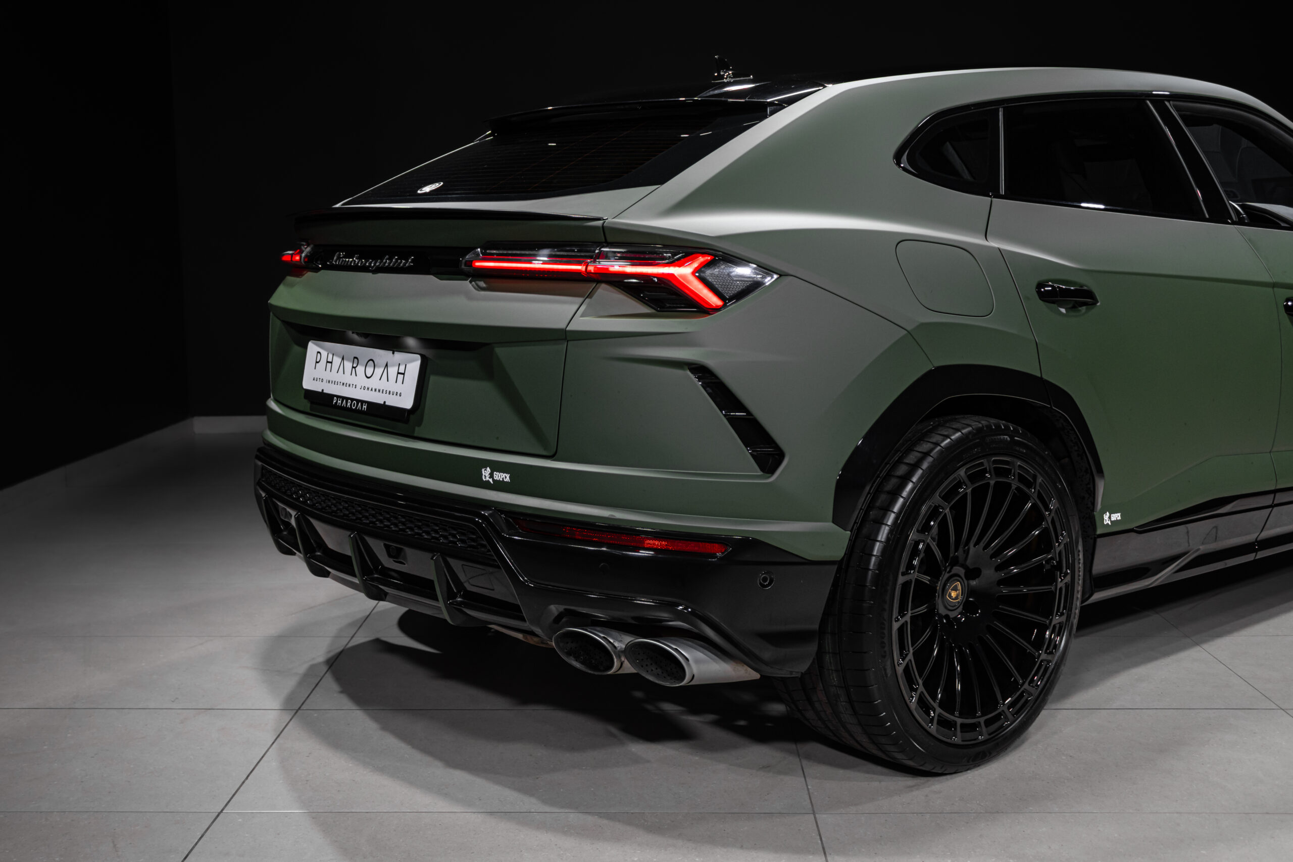 Rear three-quarter view of a matte-green Lamborghini SUV with red Y-shaped taillights, quad exhausts, and large black wheels