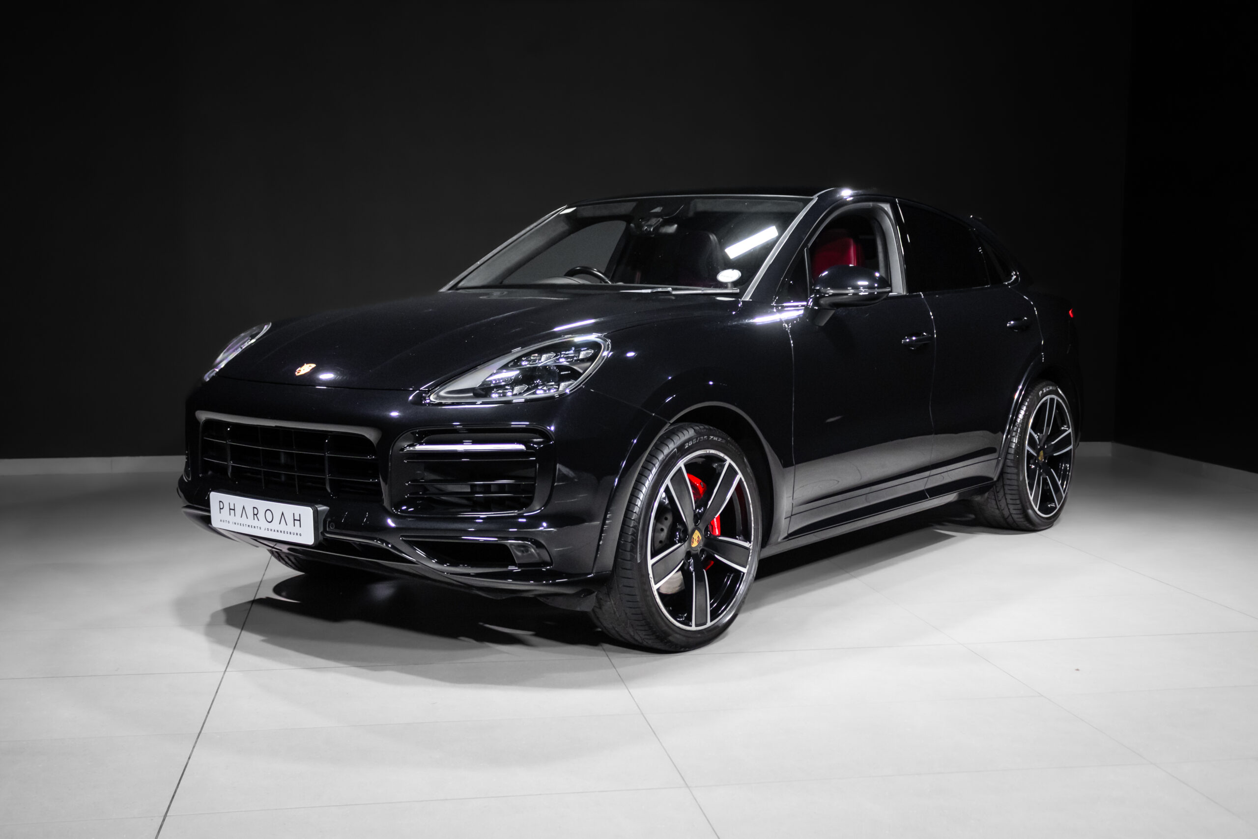 Front three-quarter view of a black luxury SUV (Porsche) in a studio showroom with red brake calipers visible through the whe