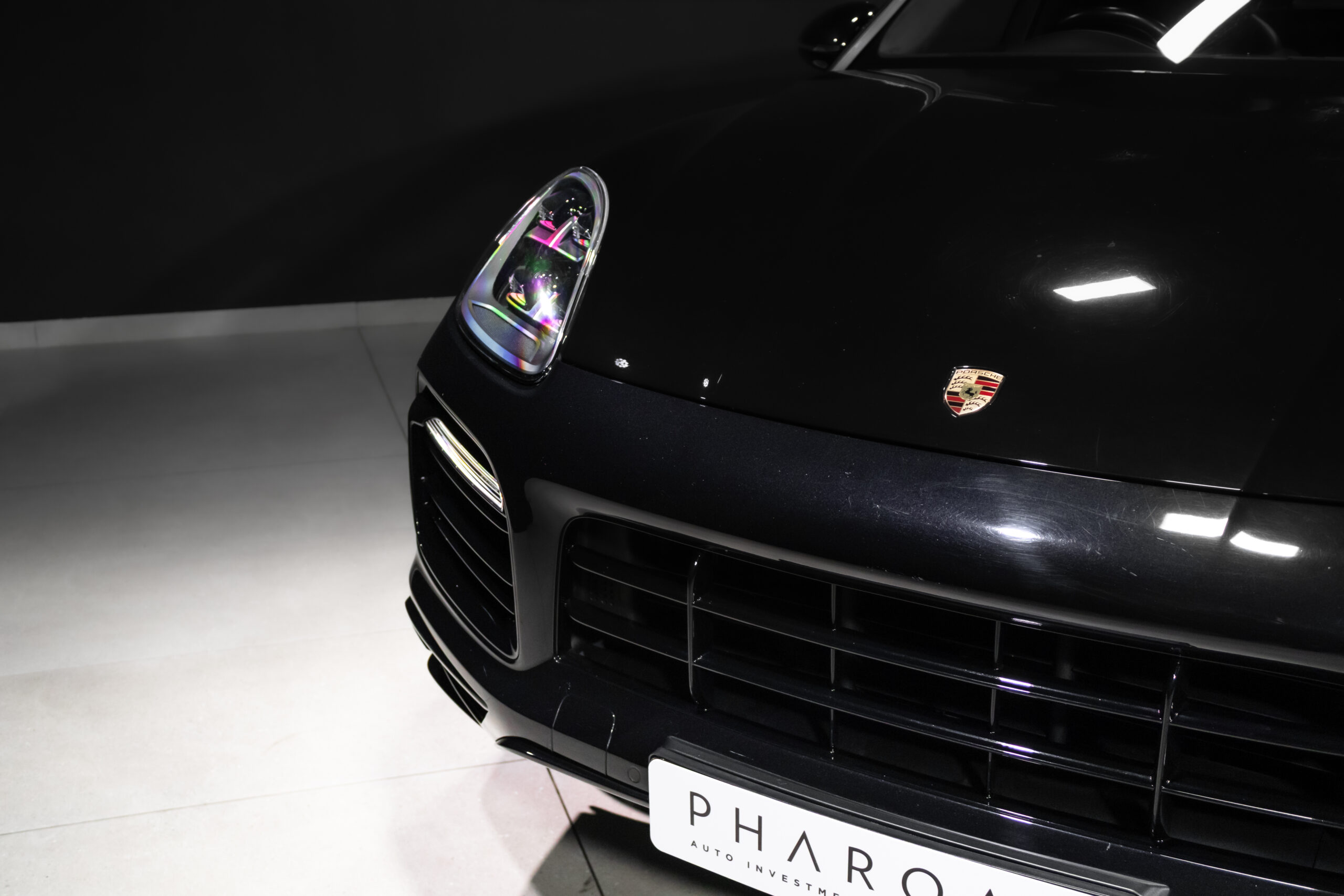 Close-up of the front left of a black Porsche, featuring the headlight, crest, and grille with a white PHAROS license plate