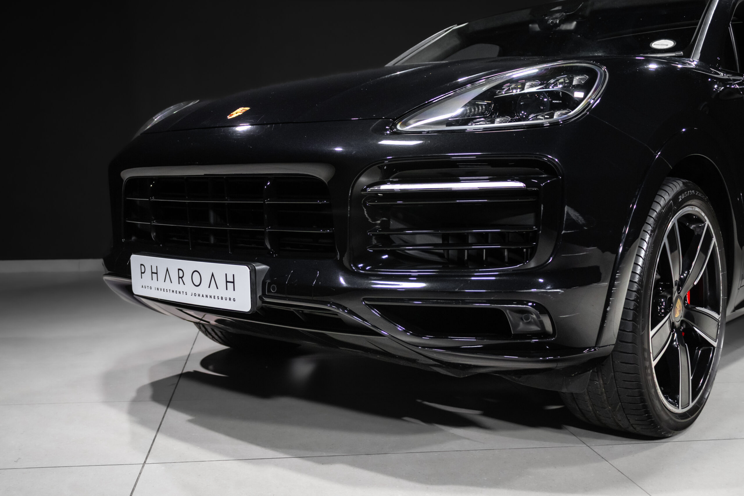 Front-right/left close-up of a glossy black Porsche in a showroom, focusing on the headlight, grille, and bumper with a 'PHAR