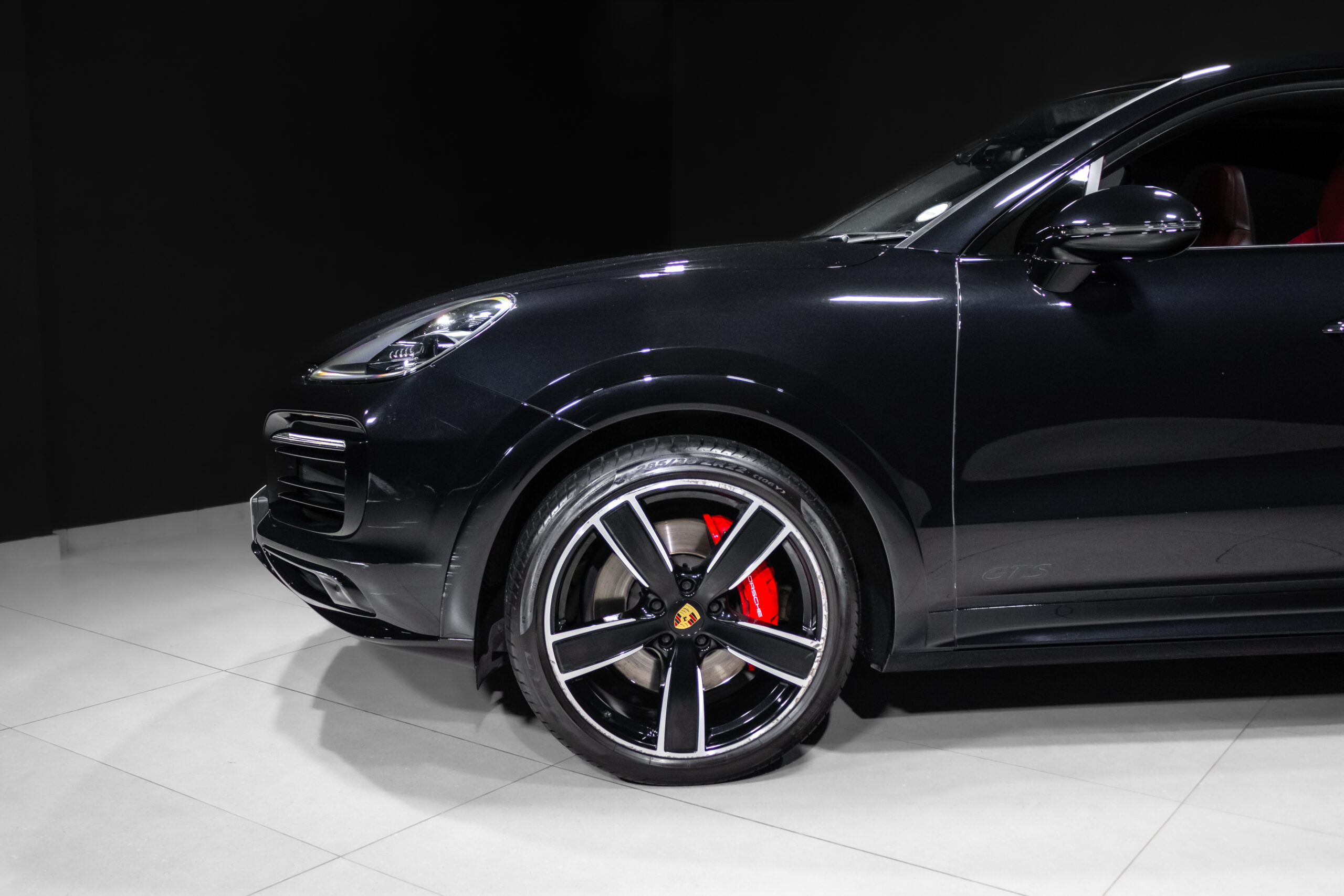 Dark Porsche sports car from the front-left side in a showroom, red brake calipers visible behind large alloy wheels