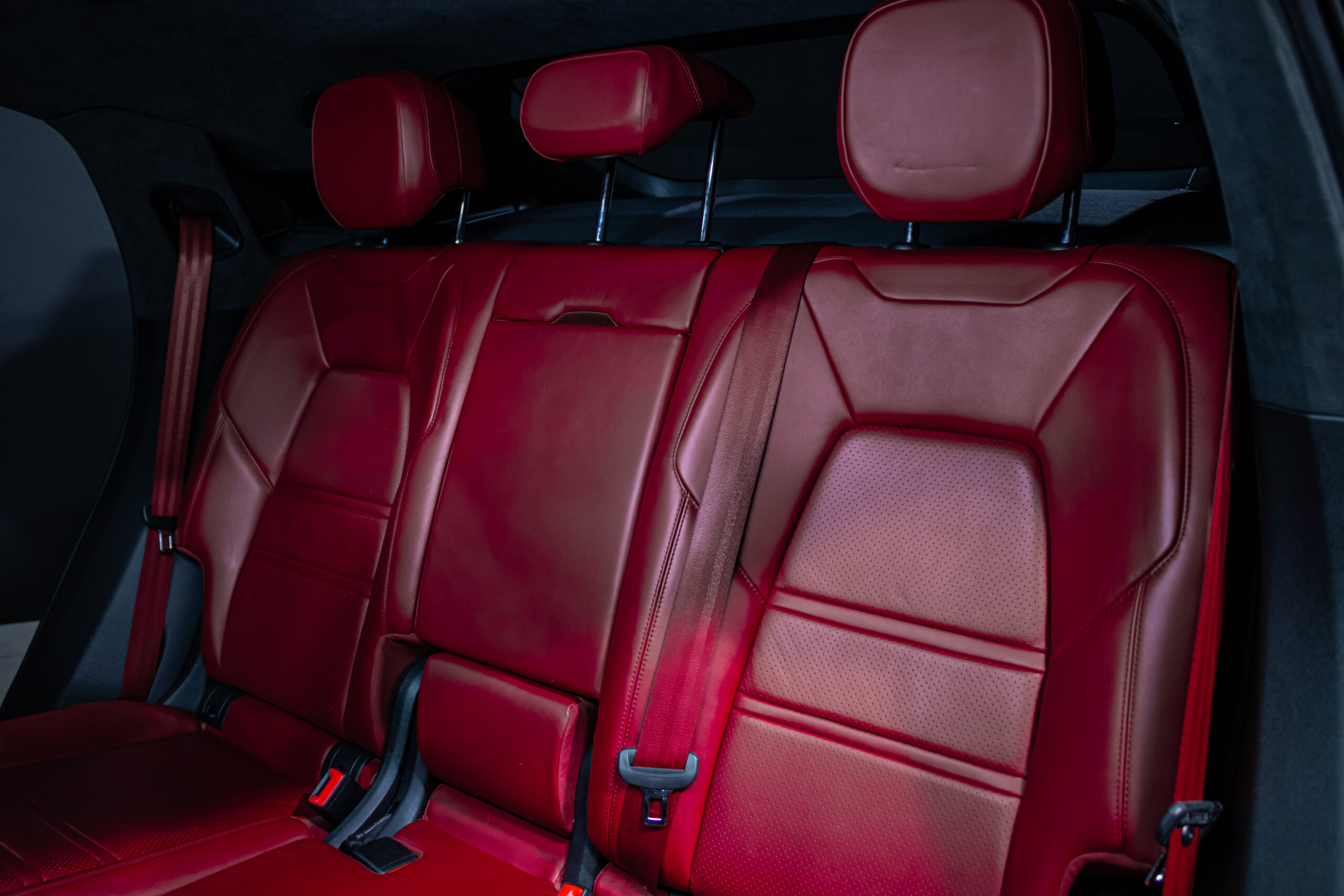 Red leather back seat row in a car with three headrests and visible seat belts, luxury interior.