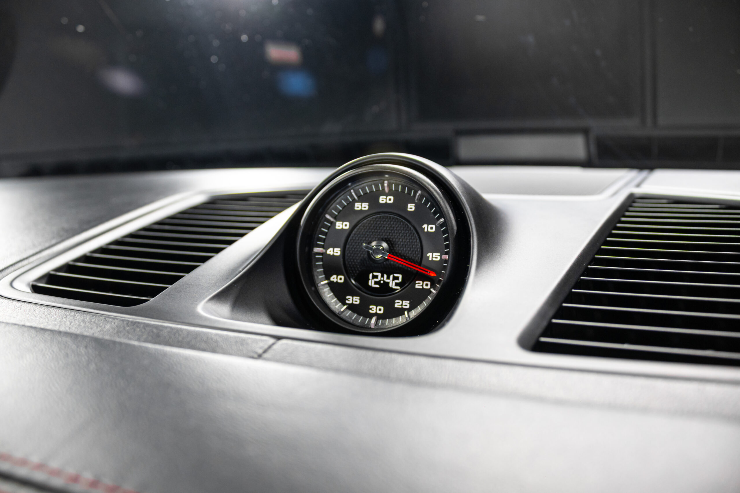 Close-up of a round car dashboard clock with a red hand and a digital display showing 12:42, mounted between the driver and p