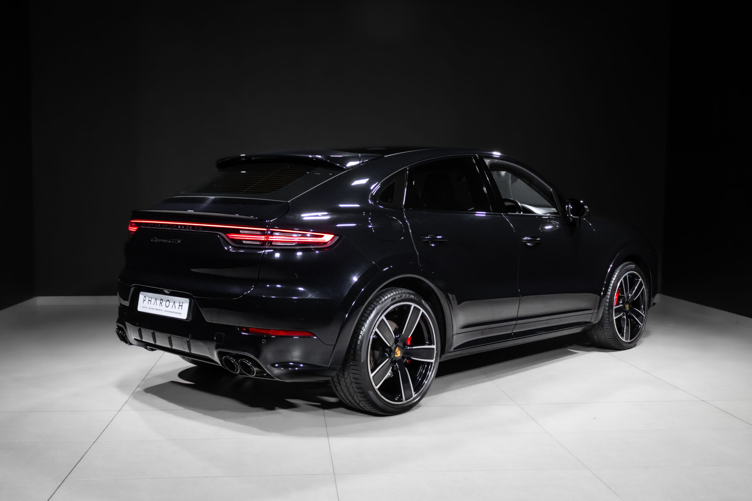 Black Porsche SUV (Cayenne) in a studio showroom, rear three-quarter view with illuminated red taillights.