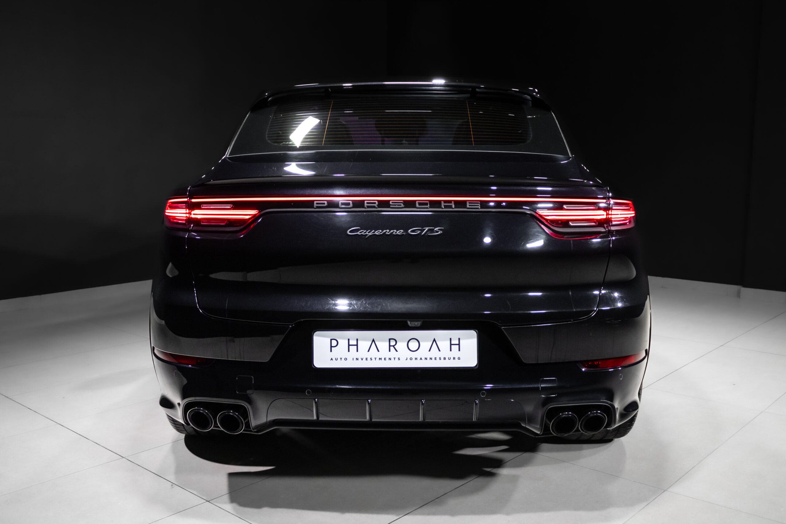 Rear view of a black Porsche Cayenne GTS in a showroom, red taillights lit and quad exhausts visible.