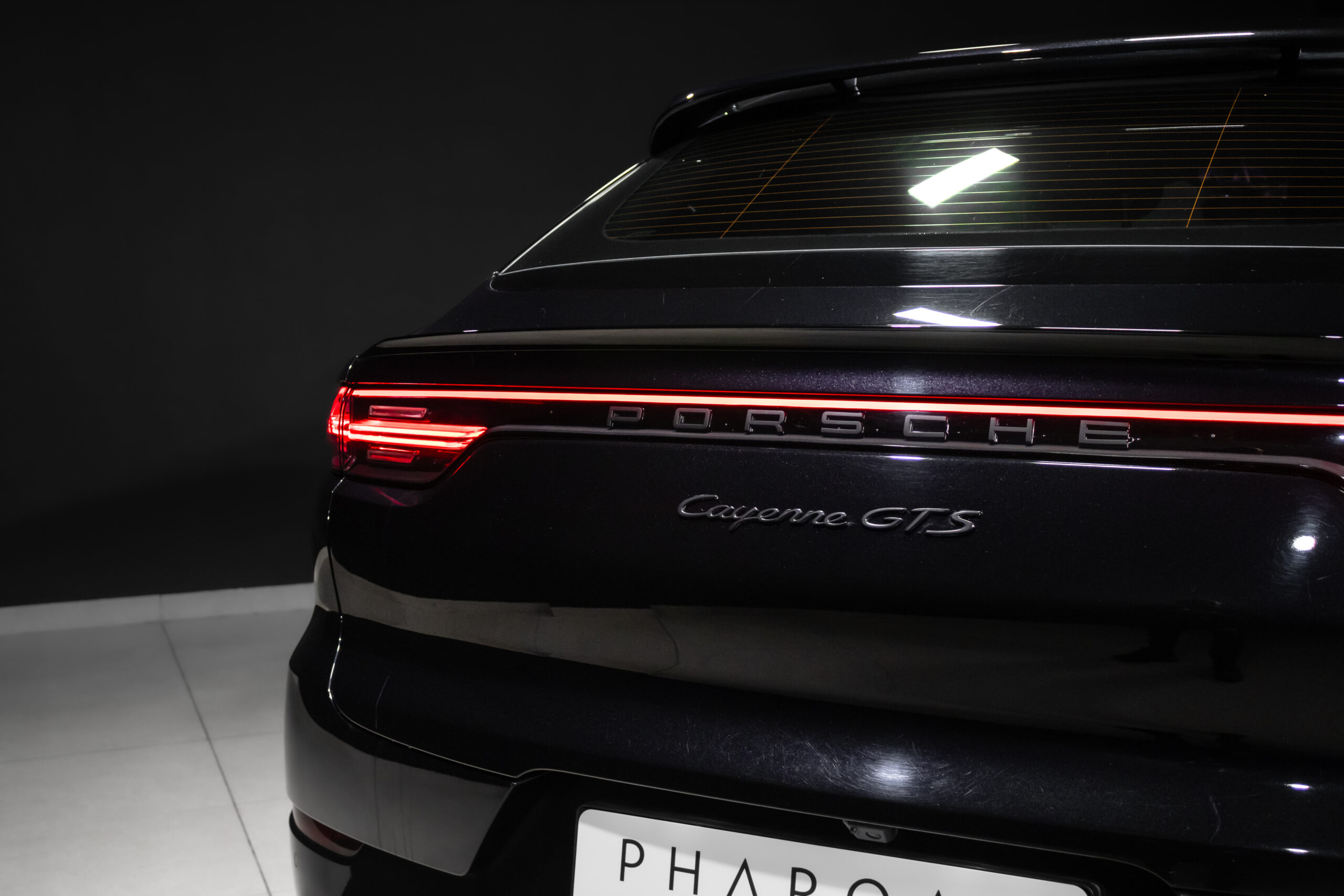 Rear view of a black Porsche Cayenne GTS in a dark showroom, featuring a continuous red taillight strip and 'Porsche' letteri