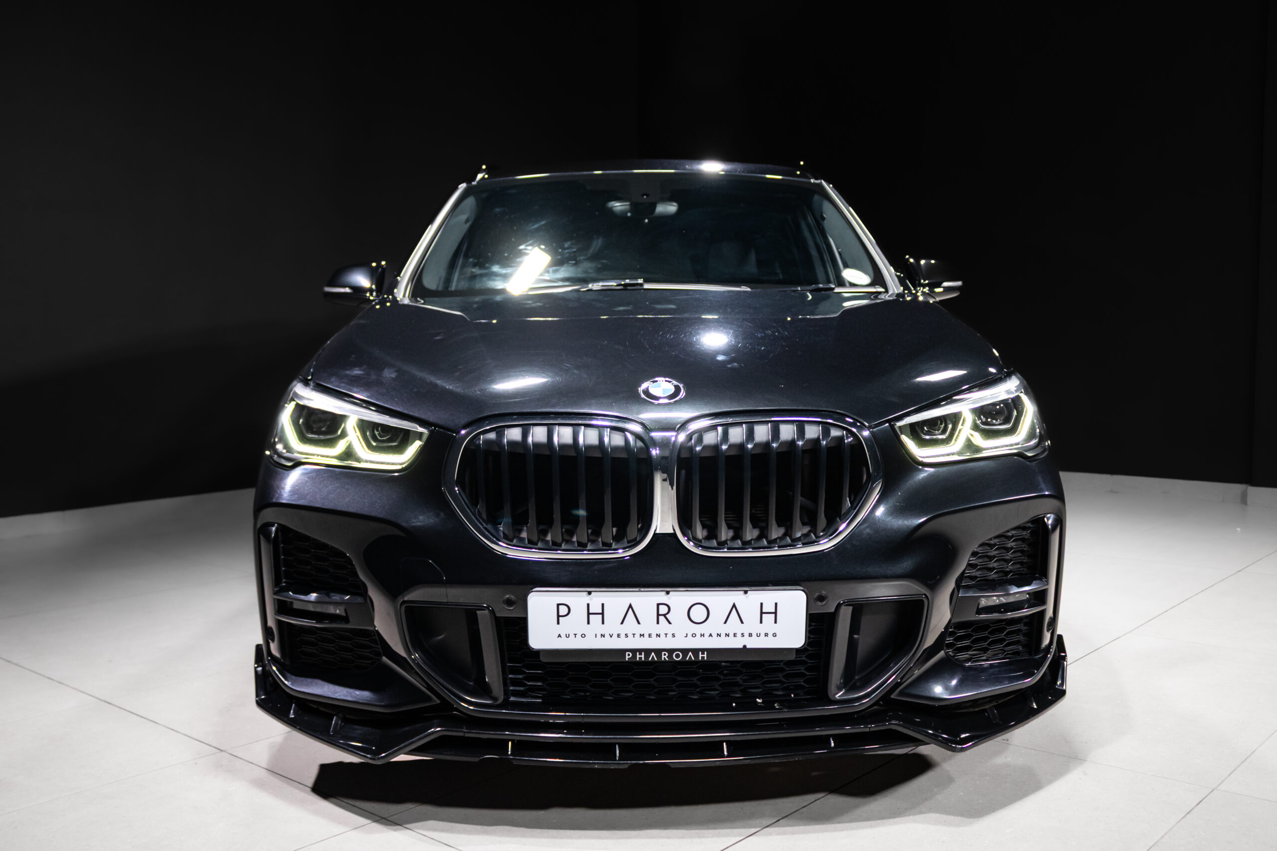 Front view of a dark BMW SUV in a showroom, illuminated headlights highlighting the iconic kidney grille and low front splitt