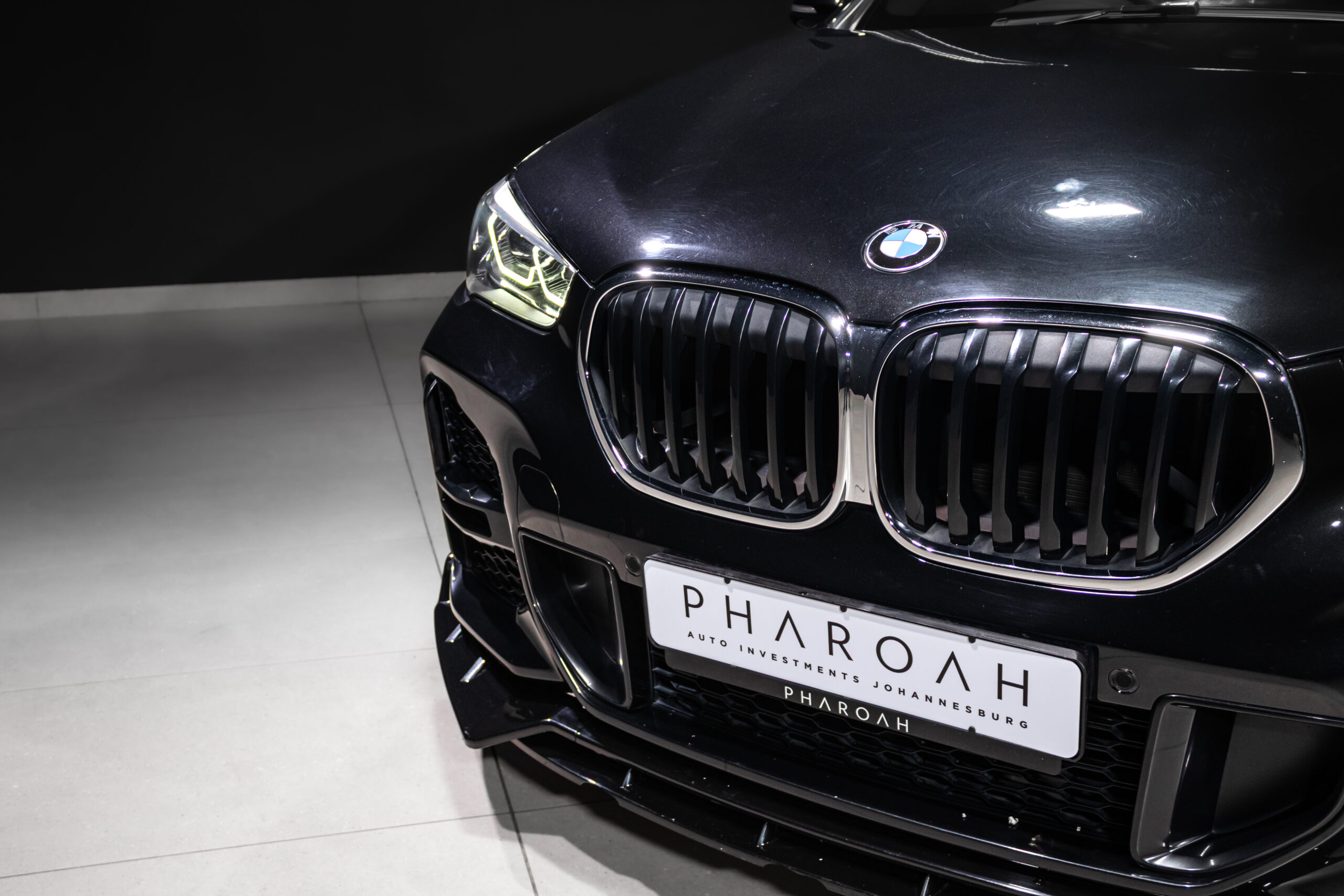 Front view of a black BMW with large kidney grille, headlight on, in a showroom; license plate reads PHAROH (auto investments