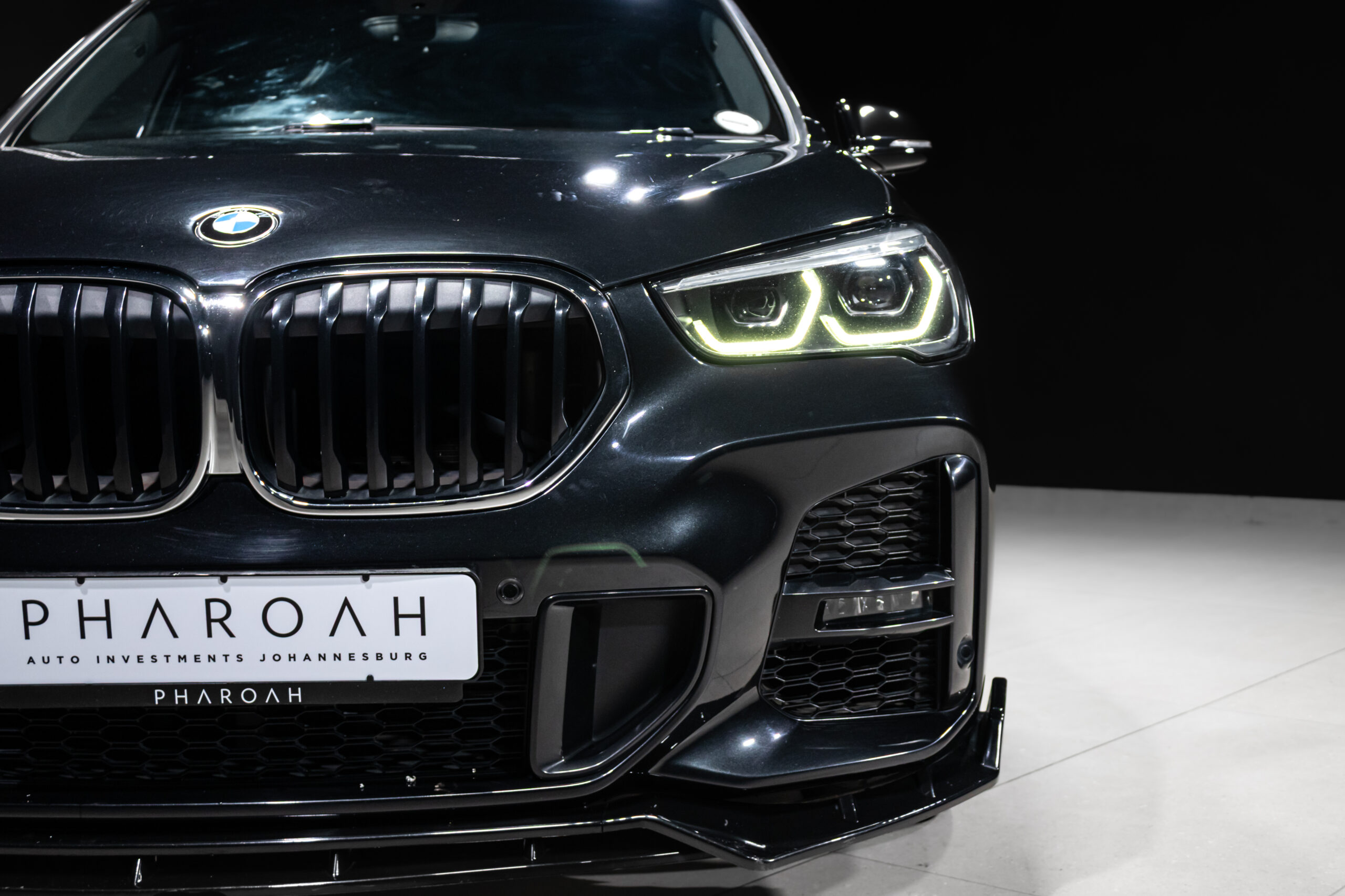 Front view of a dark BMW with illuminated twin-LED headlights and prominent kidney grille, showing a custom PHAROAH license p