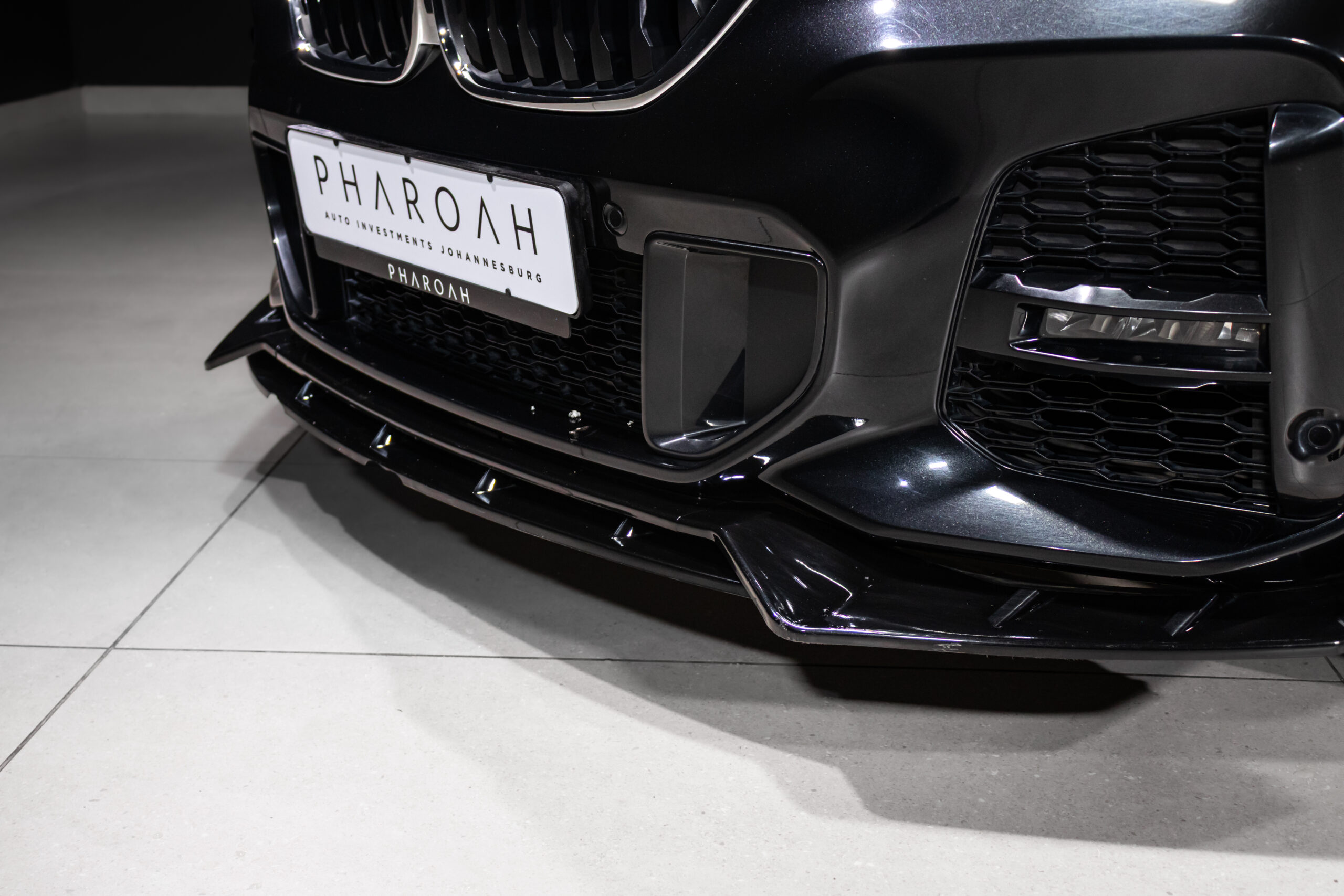 Front end of a black car with a custom Pharoah license plate and a glossy grille, bumper, and splitter visible in a showroom.