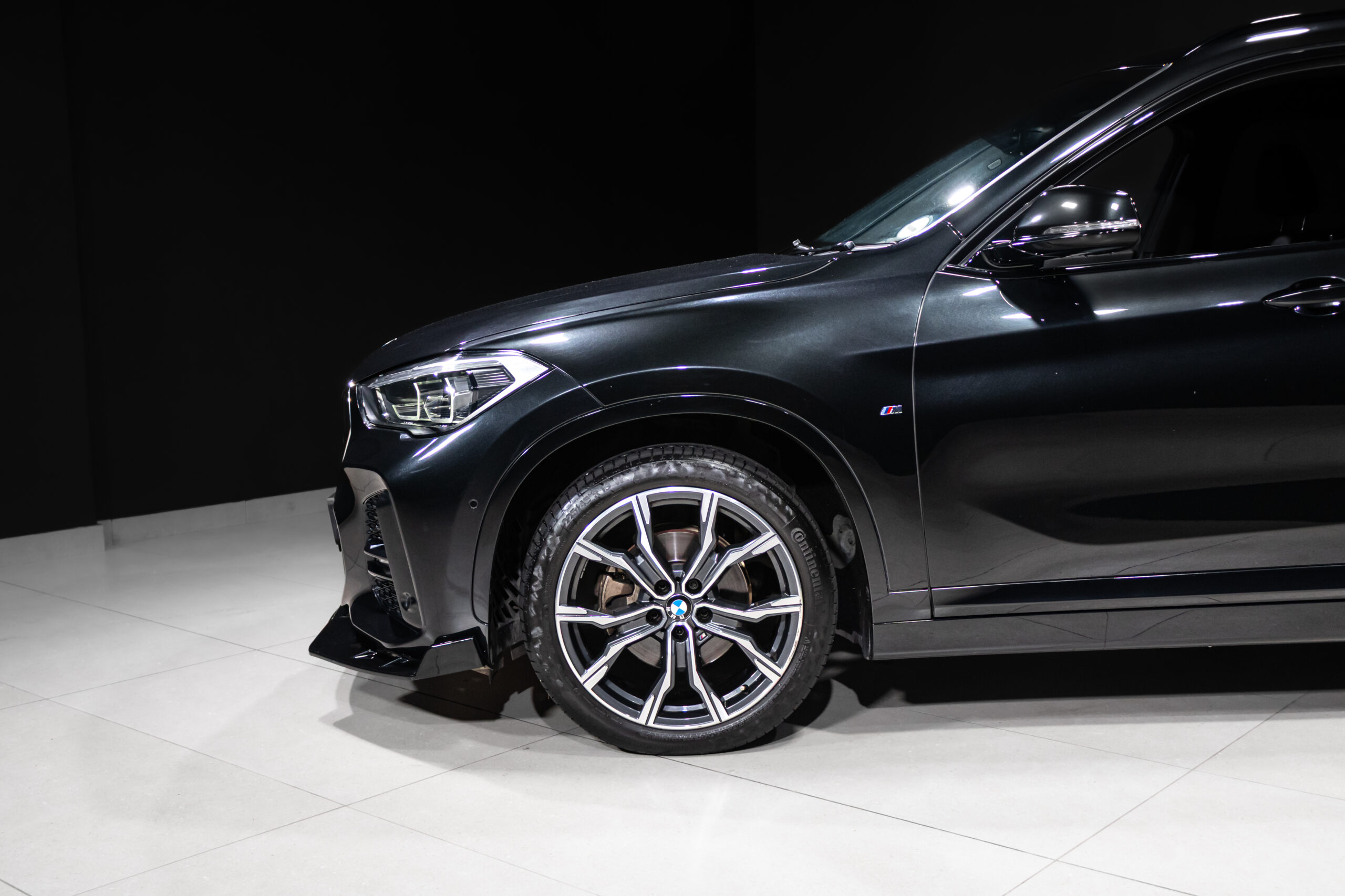 Close-up of the front left corner of a dark BMW SUV in a showroom, showing the headlight, fender with M badge, and large allo