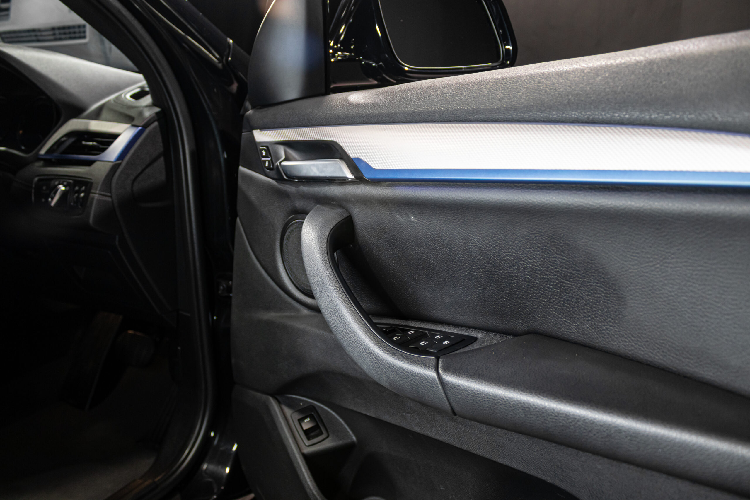 Interior of a car door panel with the armrest, window controls, and speaker visible near the front passenger seat edge.