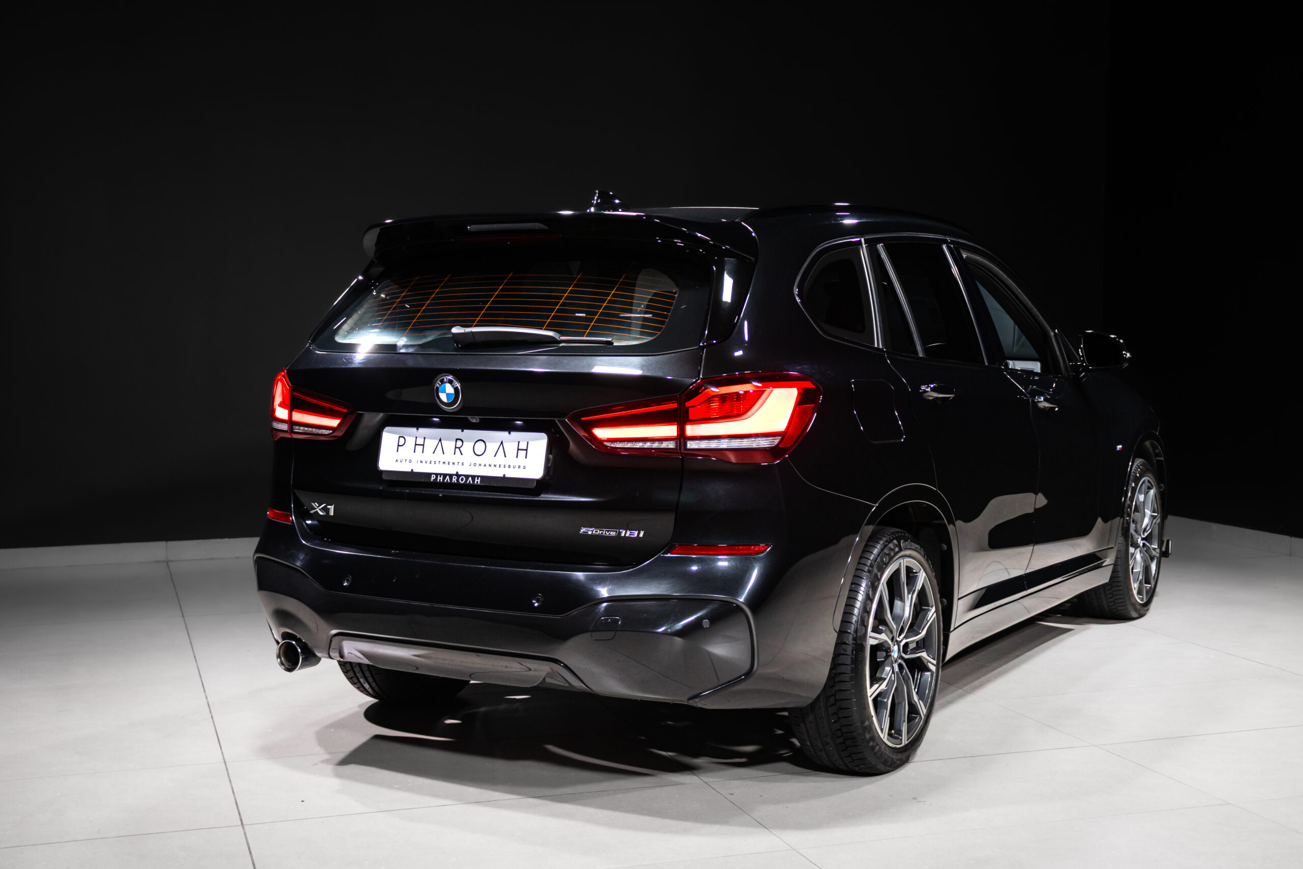Rear three-quarter view of a black BMW X1 SUV in a studio, with illuminated red taillights and a PHAROH license plate visible