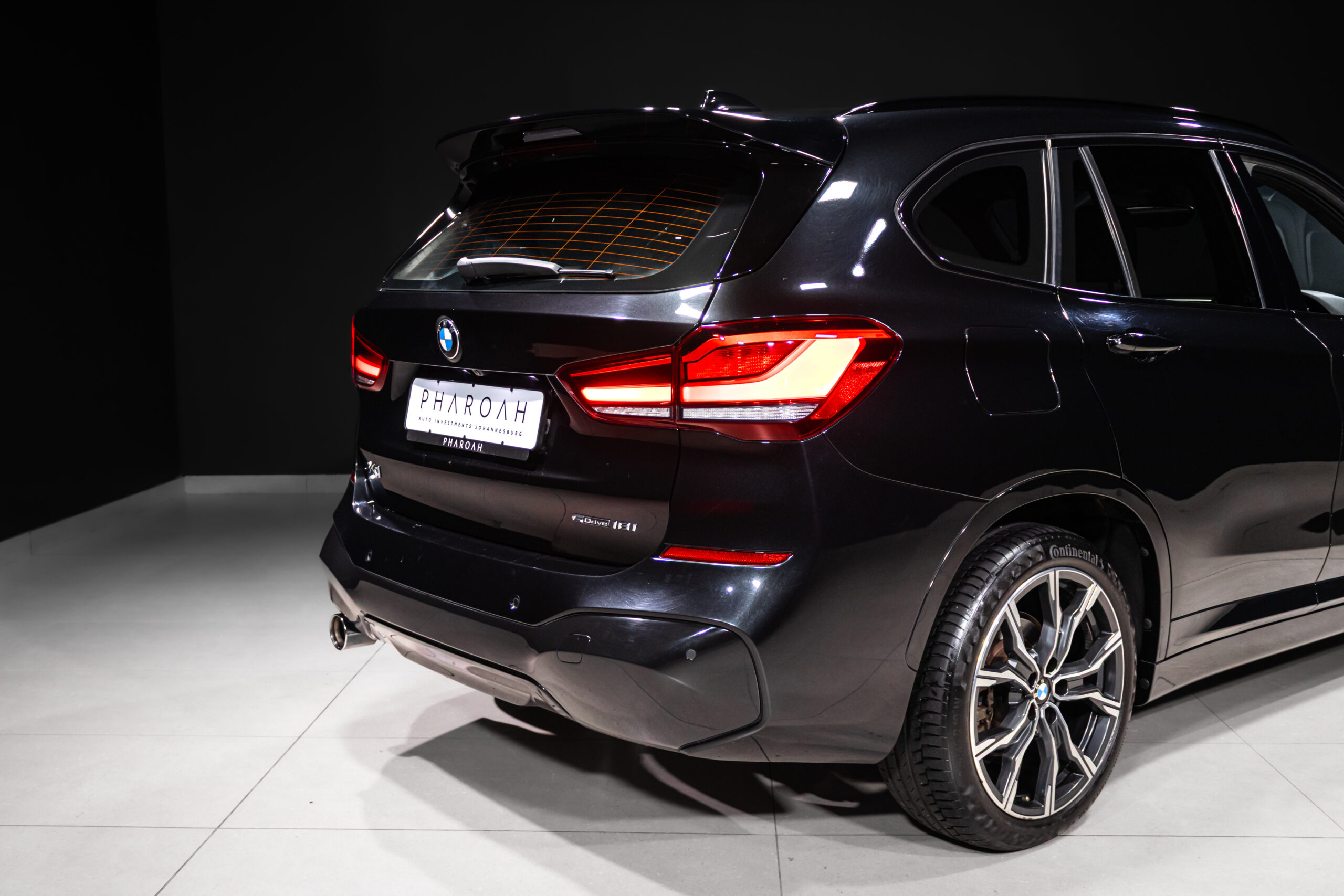 Rear-right view of a dark BMW SUV in a showroom, illuminated red taillights and glossy bodywork visible. license plate reads