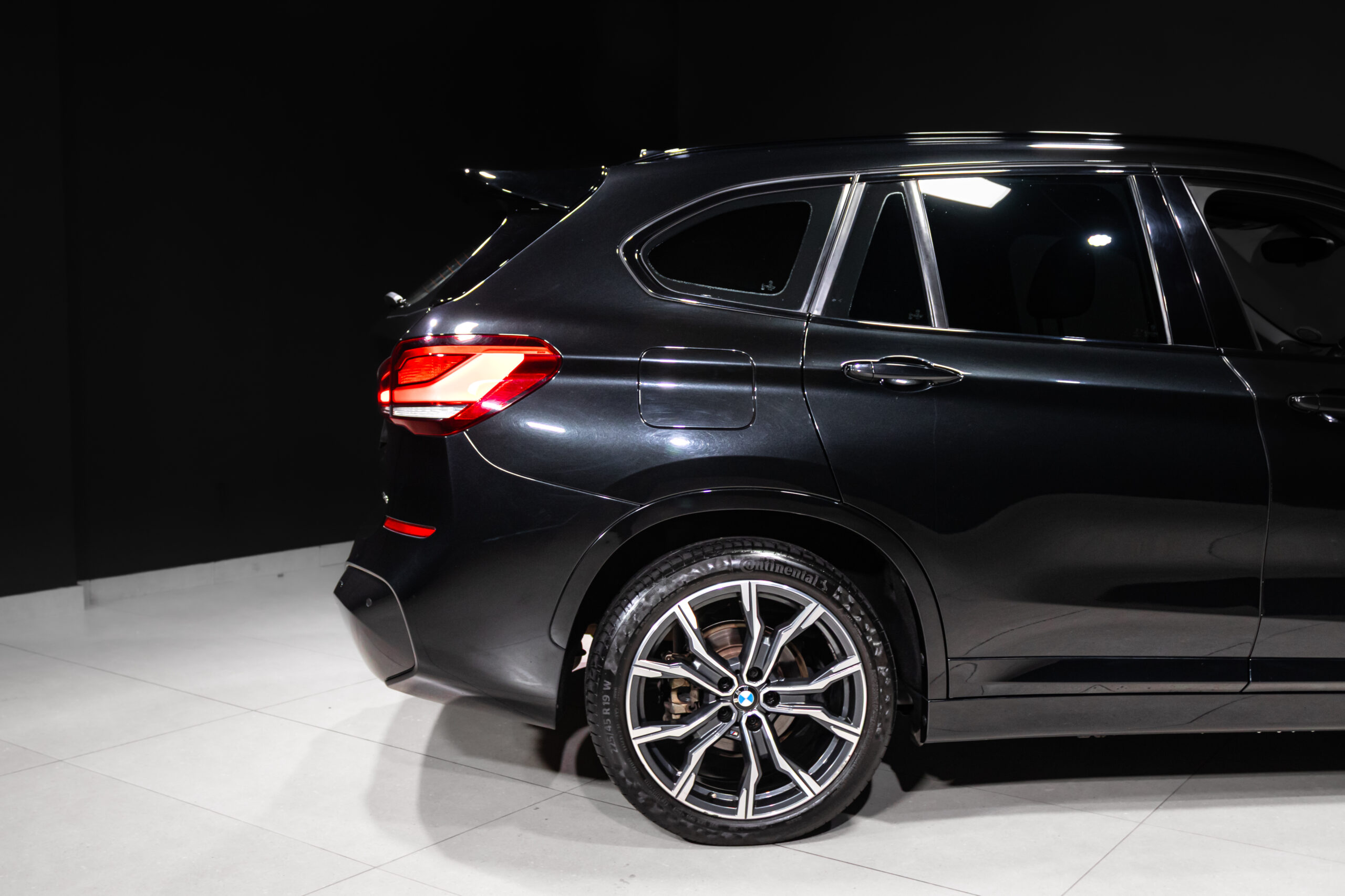 Rear quarter view of a dark gray BMW SUV in a studio, showing the illuminated taillight and stylish alloy wheel on the left r